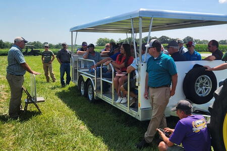 The new people mover makes field days more effective A group of people sit on an open-sided trailer listening to a man speaking at a field day